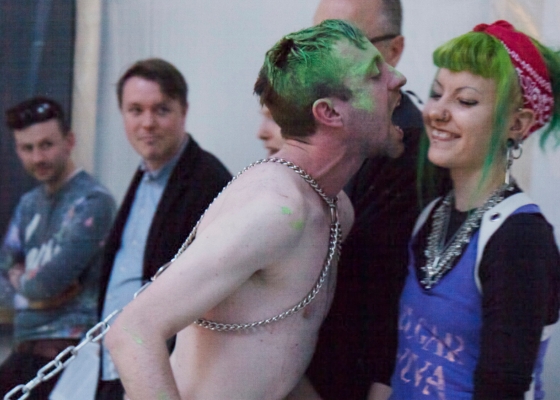 Naked man on a chain leans towards laughing woman as if to bit her